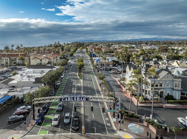 Luftbild der Hauptstraße von Carlsbad mit Ortsschild über der Fahrbahn, Palmen und Wohnhäusern, im Hintergrund der Pazifik