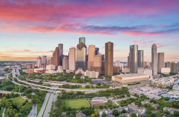 Die Hochhäuser von Houston unter rosa Abendwolken, davor Autobahnschleifen und Grünflächen