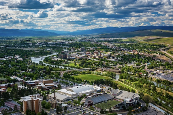 Luftbild einer Stadt im Tal mit Fluss, Bruecken und Sportstadion, dahinter bewaldete Bergketten