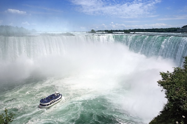 Die Niagarafälle mit aufsteigender Gischt, davor ein Ausflugsboot auf dem Fluss