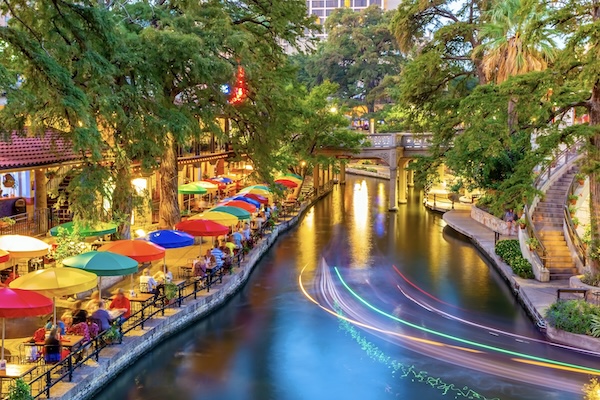 Bunte Sonnenschirme am Kanal des River Walk in San Antonio, auf dem Wasser die Lichtspur eines Boots